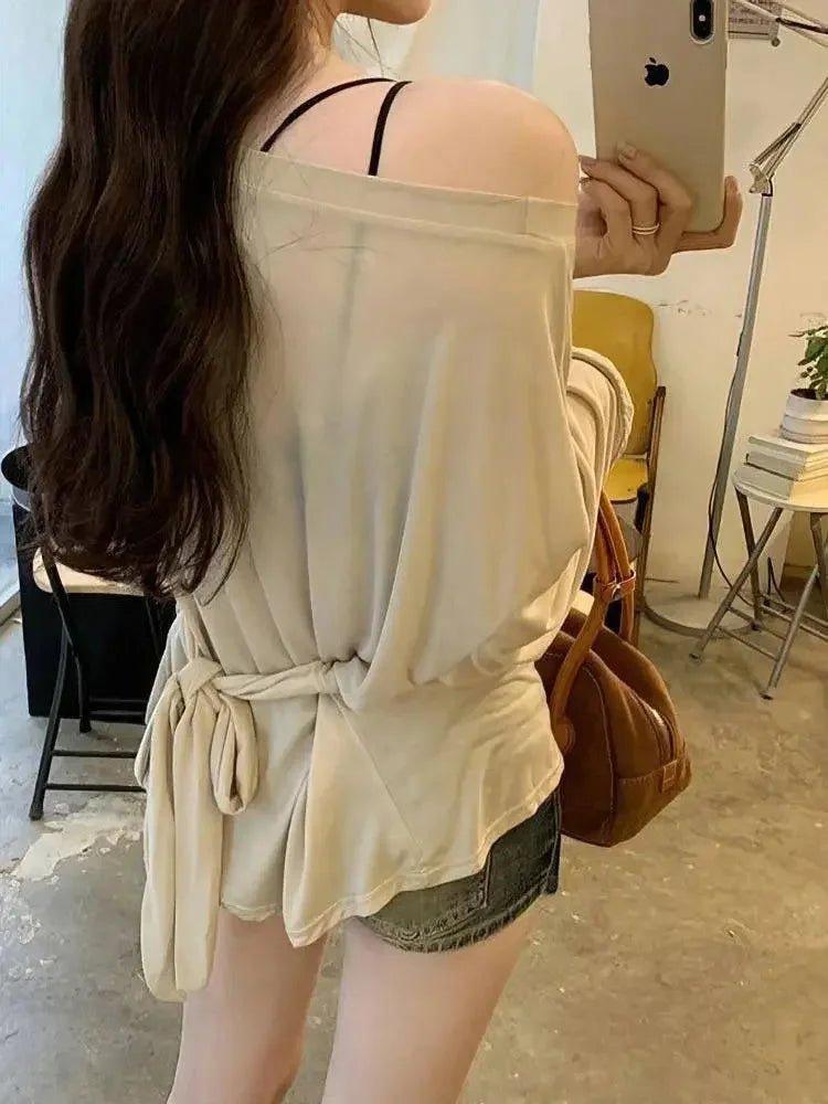 Woman wearing off-shoulder beige top with tie waist, denim shorts, taking mirror selfie