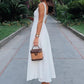 Woman in elegant white midi dress with back cutout, holding woven handbag, walking garden path