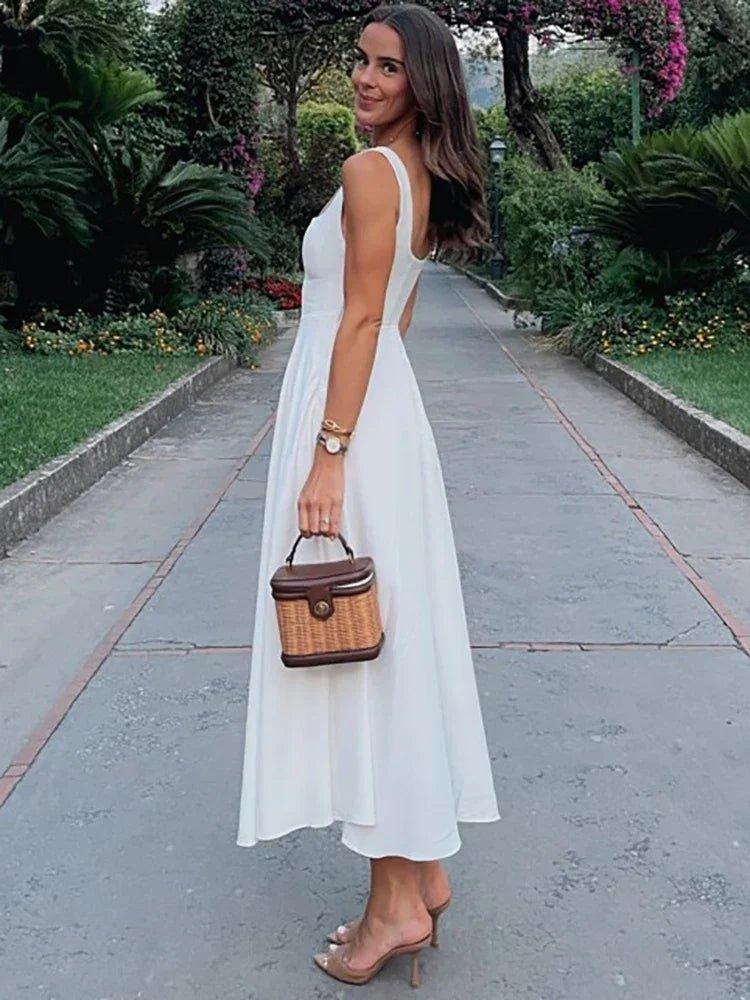 Woman in elegant white midi dress with back cutout, holding woven handbag, walking garden path