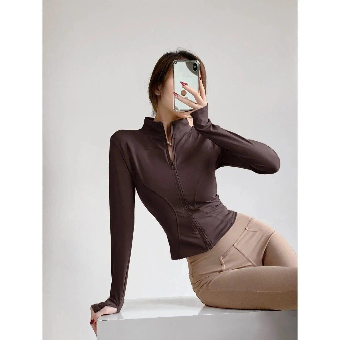 Woman in brown zip-up athletic top and beige leggings posing with phone