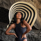Woman in black swimsuit wearing large striped sun hat against rocky backdrop