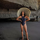 Woman in navy one-piece swimsuit and striped sun hat on black sand beach under rock formation