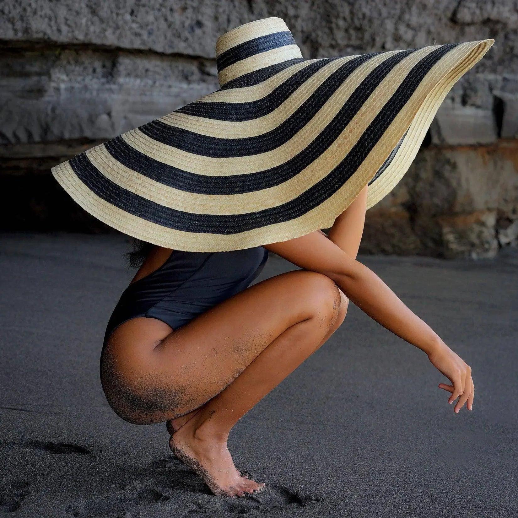 Woman in black swimsuit crouching on beach with oversized striped sun hat