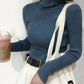 Woman wearing blue ribbed turtleneck sweater with white overalls and black belt, holding pink drink