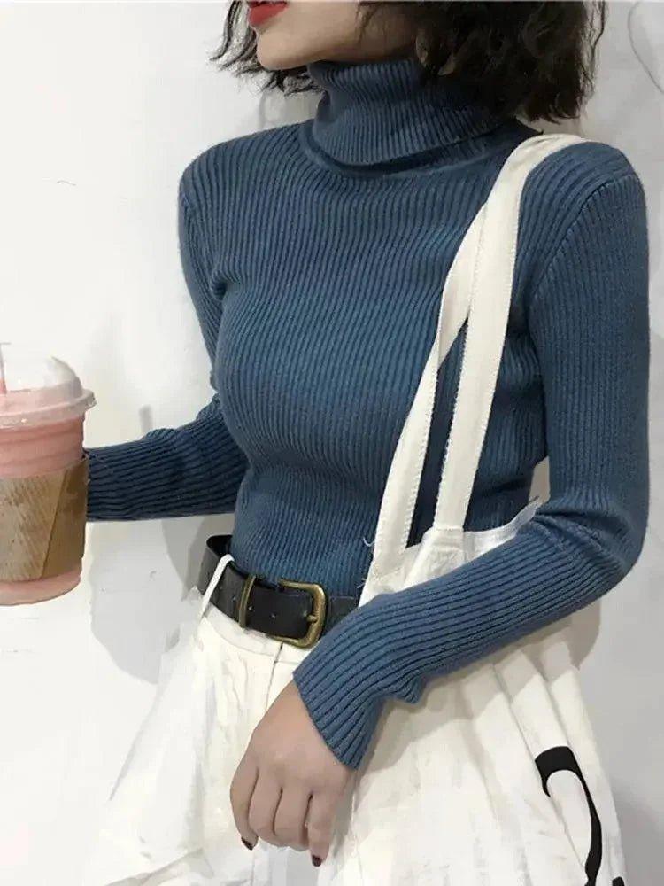 Woman wearing blue ribbed turtleneck sweater with white overalls and black belt, holding pink drink