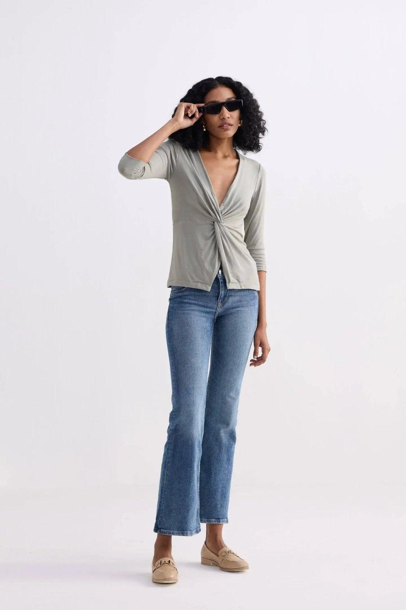 Woman in stylish green twist-front top, blue jeans, and loafers, adjusting sunglasses for chic fashion look