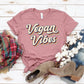 Pink 'Vegan Vibes' graphic tee with plaid scarf, denim, cotton stems, and suede boots on wood