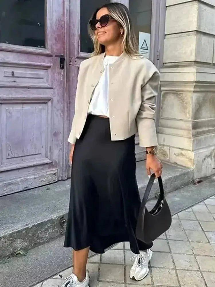 Woman in beige jacket, black midi skirt, white sneakers, and sunglasses, holding black handbag