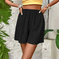 Black pleated women's shorts with elastic waist, styled with yellow top and bracelets, tropical leaf backdrop
