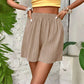 Beige pleated women's shorts with elastic waist, paired with yellow crop top, tropical leaf backdrop