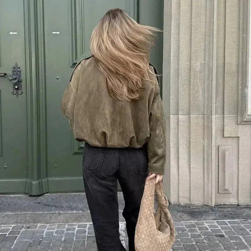 Woman in olive suede jacket and black jeans, holding woven bag, walking past green door — stylish street fashion